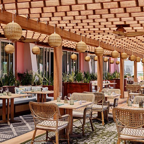 An outdoor restaurant features wicker chairs, wooden tables, and hanging lanterns under a pergola, creating a cozy and inviting atmosphere.
