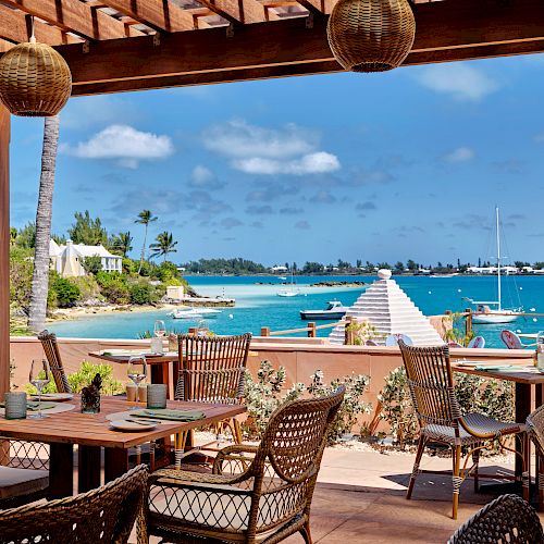 An outdoor dining area with wicker chairs and wooden tables overlooks a coastal view with boats on a bright, sunny day and a clear blue sky.