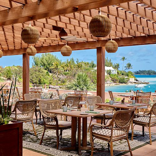 An outdoor restaurant with wicker furniture under a pergola, overlooking a scenic view of greenery and water with boats in the distance.