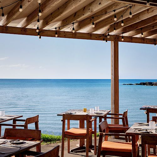 An outdoor seaside restaurant with wooden tables and chairs under a pergola, overlooking a calm ocean view.