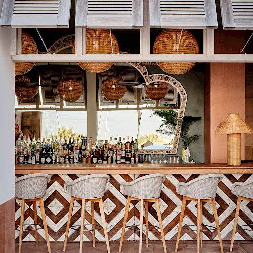 A stylish bar features a chevron-patterned counter, four stools, hanging wicker lights, and a variety of bottles in front of a large mirror.