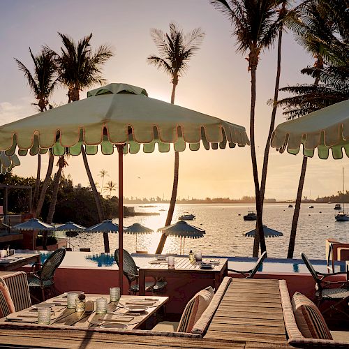 A serene beachfront scene with tables and chairs under green umbrellas, surrounded by palm trees, facing the calm sea at sunset with boats in the distance.
