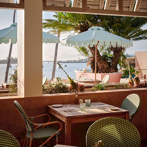 An outdoor dining area with light green chairs and tables is beneath umbrellas, overlooking a beach setting with palm trees and ocean views.