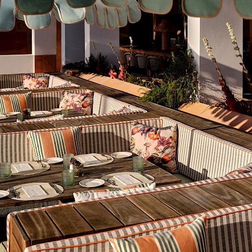 An outdoor dining area with wooden tables, striped cushioned seating, and floral throw pillows under a wavy green canopy. Plates and utensils are set.