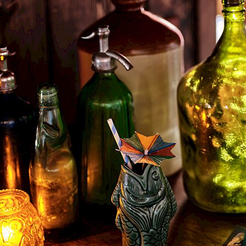 The image features various vintage bottles, a tiki-style ceramic mug with a straw and umbrella, a lit candle, and a matchbox on a wooden surface.
