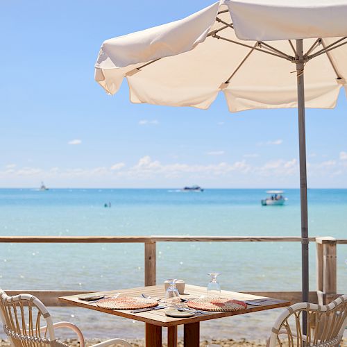 A seaside dining setup with a table, two chairs, an umbrella, and a view of the ocean under a clear blue sky, ending the sentence.