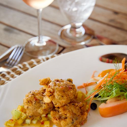 A gourmet meal consisting of what appears to be crab cakes, a side salad, and garnishes, served with a glass of wine and water, on a wooden table.