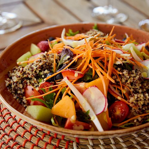A colorful salad with mixed greens, quinoa, shredded carrots, radishes, cherry tomatoes, and other vegetables in a ceramic bowl on a woven placemat.