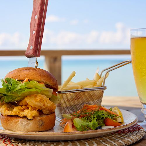 A gourmet burger with a knife through it, a basket of fries, a small salad, and a glass of beer on a table by the seaside.