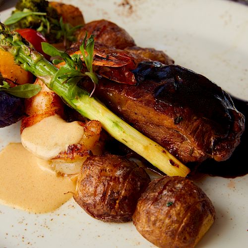 A plate of gourmet food featuring a meat dish with sauce, roasted potatoes, asparagus, colorful vegetables, and a creamy sauce.
