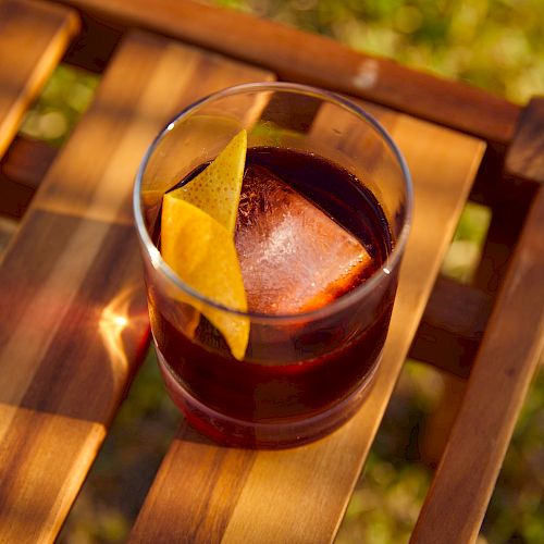 A glass of dark-colored beverage with a large ice cube and a lemon twist garnish rests on a wooden surface outdoors, with sunlight casting shadows.