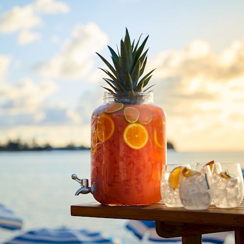 A large glass jar filled with a red punch, topped with a pineapple, and two glasses with drinks on a wooden table by the seaside at sunset.