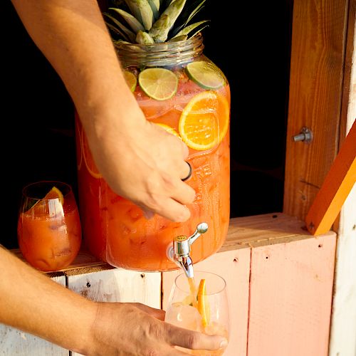 A person is dispensing a tropical fruit drink from a large jar with a tap into a glass. The jar is decorated with citrus slices and a pineapple top.