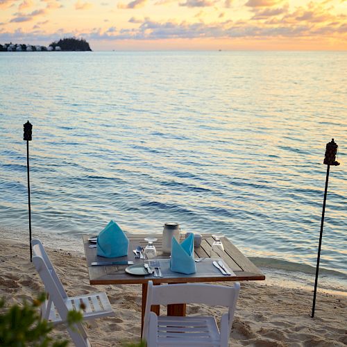 A beachfront dining setup with two chairs and a table set for two, featuring blue napkins, surrounded by torches, overlooking a serene ocean at sunset.