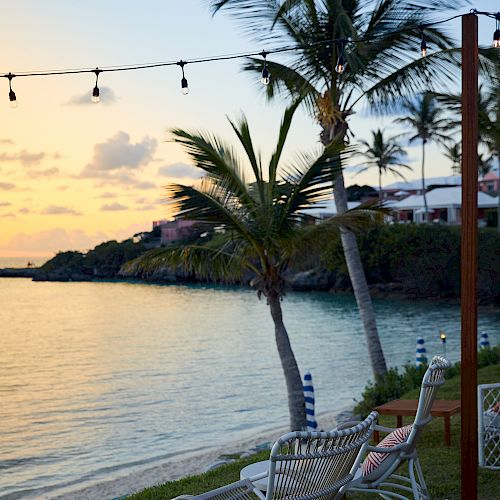 The image shows a serene beach scene at sunset with two chairs, palm trees, a string of lights, and calm water.