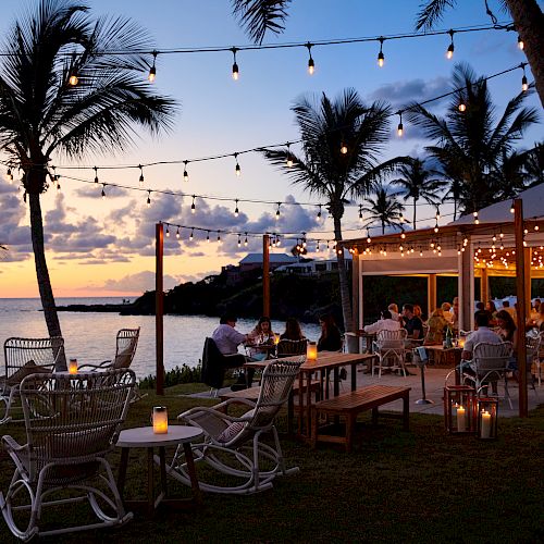 An outdoor beachside restaurant at sunset, decorated with string lights and palm trees. People dine and relax, creating a cozy atmosphere.