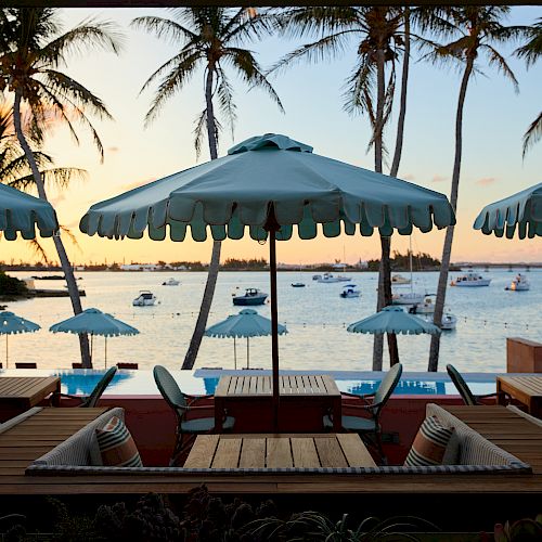 A scenic beachfront view with tables and umbrellas by a deck, boats on the water, and palm trees silhouetted against a serene sunset.