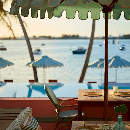 A serene seaside view with tables, chairs under an umbrella, plates, glasses, and boats on the water in the background in the evening.
