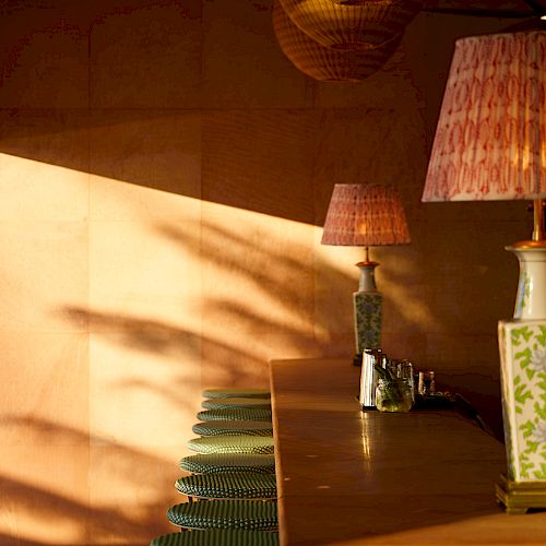 A cozy indoor bar setting with stools, warm lighting, and lamps casting shadows on the wall. The decor features wicker and wooden elements.