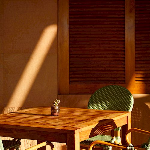 A wooden table with four wicker chairs is placed against a window with closed wooden shutters, bathed in warm sunlight.