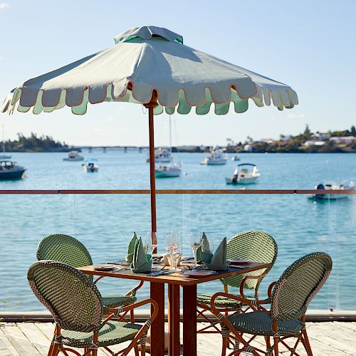 A dining table with four chairs under a large umbrella is set on a dock, overlooking a tranquil water scene with boats in the background.