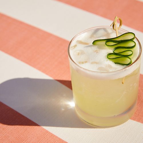 A glass of a cocktail with a frothy top, garnished with cucumber slices, set on a striped surface with shadows cast by the sunlight.