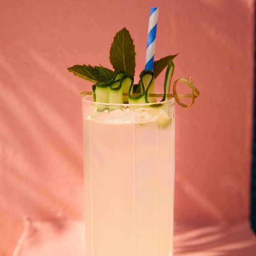 A tall glass of light-colored beverage garnished with mint leaves, cucumber slices, and a blue-striped straw, placed against a pink background.