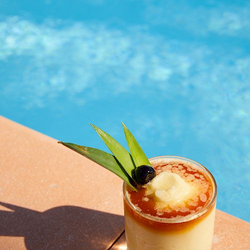 A tropical cocktail garnished with pineapple leaves and a cherry sits by the edge of a swimming pool with clear blue water in the background.