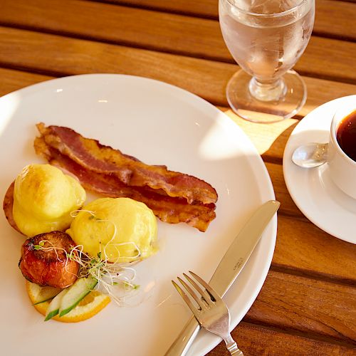 The image shows a plate with bacon, eggs Benedict, tomatoes, and garnishes beside a glass of water and a cup of coffee on a wooden table.