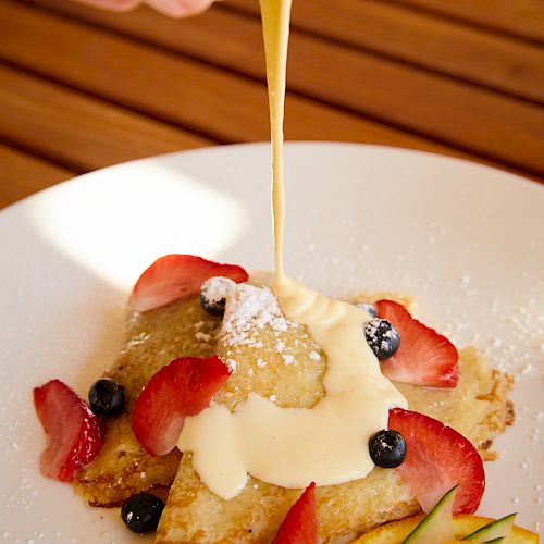A person is pouring sauce over pancakes garnished with strawberries, blueberries, and powdered sugar, served with sliced fruit on a white plate.
