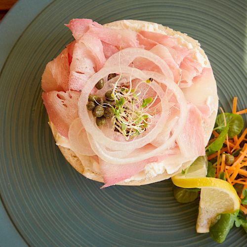 The image shows an open-faced sandwich with sliced ham, onions, sprouts, capers, and a side salad containing lemon, carrots, and greens.