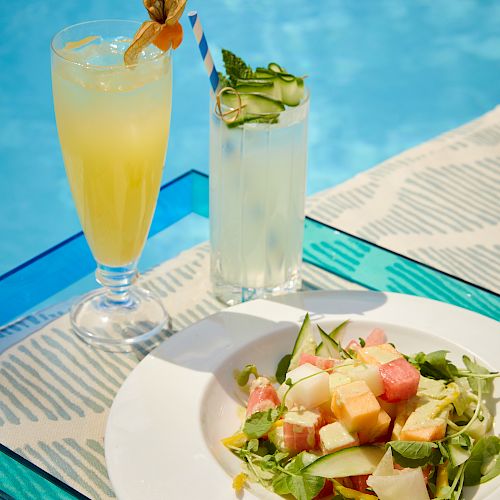 The image shows two refreshing drinks and a plate of salad placed beside a pool, creating a relaxing and summery dining scene.