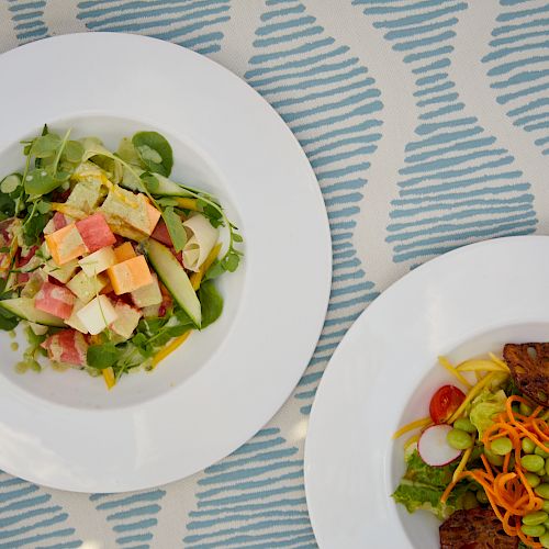 The image shows two plates of salad on a patterned tablecloth. One salad has mixed greens and diced ingredients, while the other includes sliced vegetables.