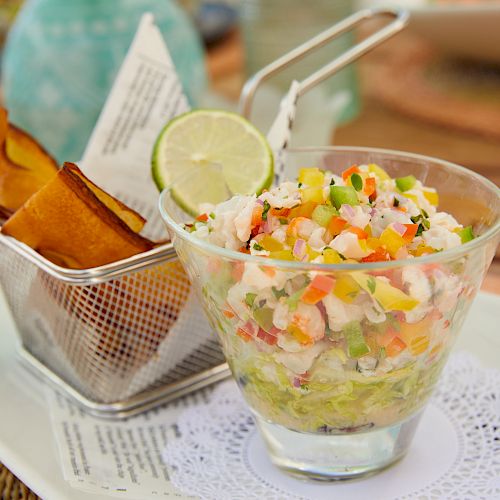 A glass of ceviche garnished with diced veggies and lime, accompanied by plantain chips served in a metal basket on a lace-lined plate.
