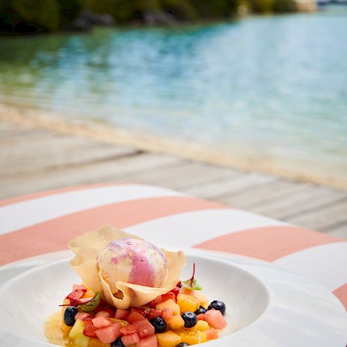 A plated dessert with fresh fruit and ice cream, set on a table by a serene water view with lush greenery and a partly cloudy sky.