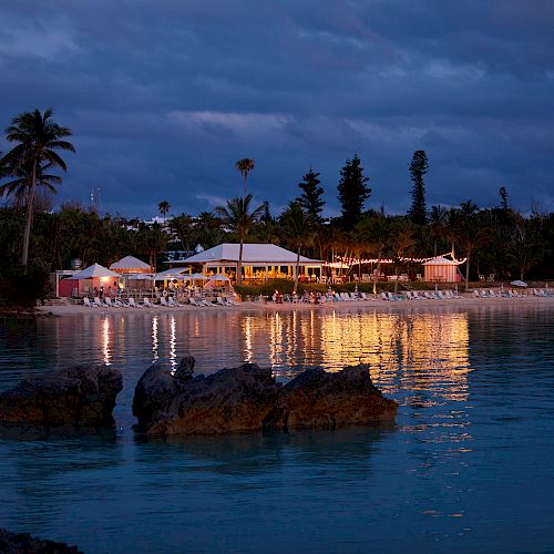 A serene beach scene at dusk, with palm trees, illuminated buildings, and their reflections in the calm water creating a picturesque view.