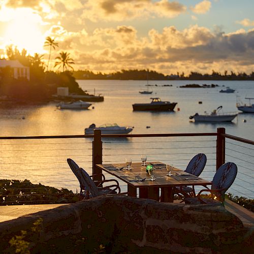 An outdoor dining setup overlooking a serene body of water with boats and a picturesque sunset in the background, framed by palm trees and foliage.