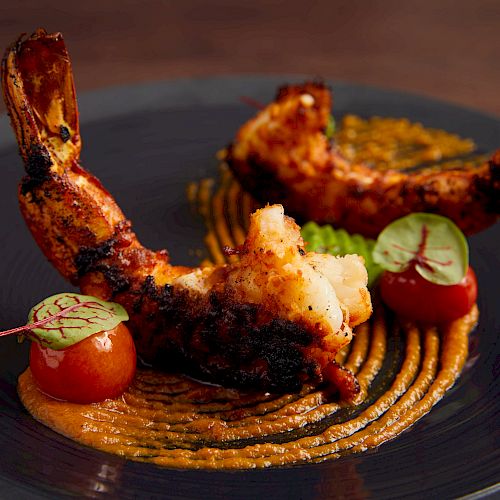 The image shows a gourmet dish with two grilled shrimp, cherry tomatoes, green garnish, and sauce artfully presented on a dark plate.