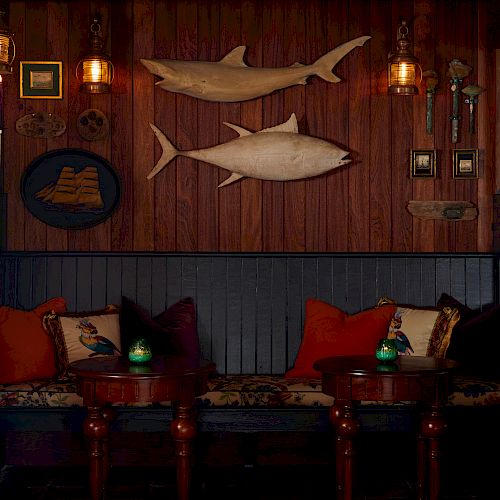 The image shows a cozy, wooden room featuring a bench with colorful pillows, two round tables, and wall-mounted decor, including two large fish.