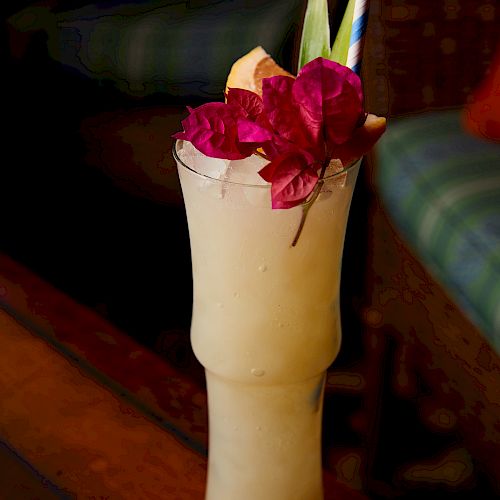 A tall glass of a creamy white beverage garnished with colorful flowers, an orange slice, and a straw, set against a blurry background.