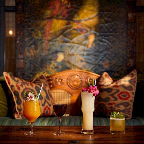 The image shows a set of four different cocktails placed on a wooden table in front of artistically arranged cushions and a decorative backdrop.