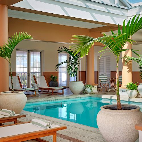 An indoor pool area with tropical plants, sun loungers, and tables. Large windows allow natural light, creating an inviting and serene atmosphere.