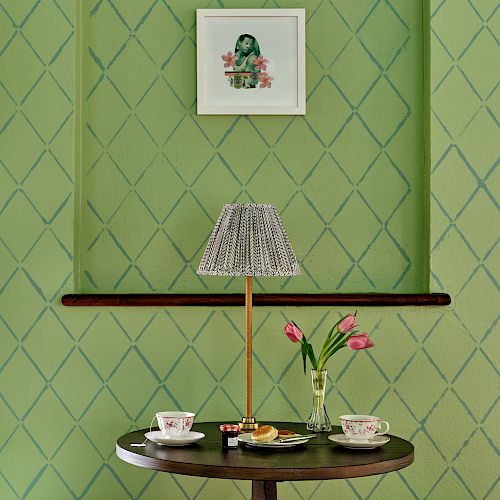 The image shows a green, patterned wall with a framed picture, a table with a lamp, teacups, a vase with pink tulips, and seats on either side.