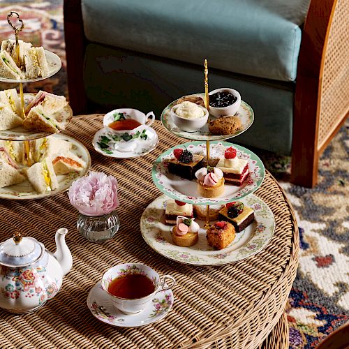 The image shows an elegant afternoon tea setup with a variety of pastries, sandwiches, a teapot, and teacups on a wicker table in a cozy seating area.