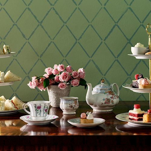 This is an elegant tea setup with a teapot, teacups, tiered trays of sandwiches and desserts, and a vase of pink roses on a wooden table with a patterned wallpaper.
