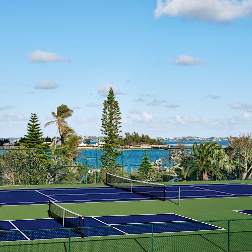 A set of blue tennis courts surrounded by green fencing, with a backdrop of trees, a large body of water, and a blue sky dotted with clouds.