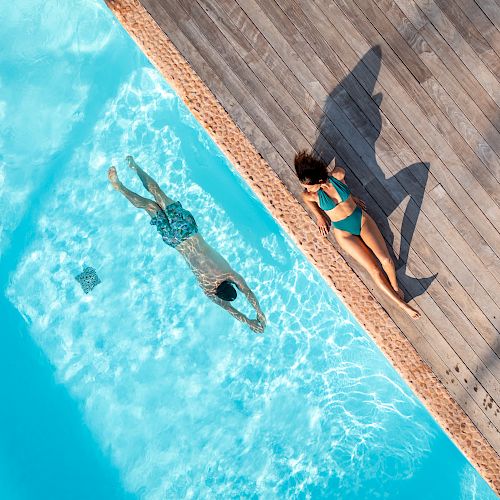 An aerial view of a swimming pool with a person swimming underwater and another person sunbathing at the pool's edge.