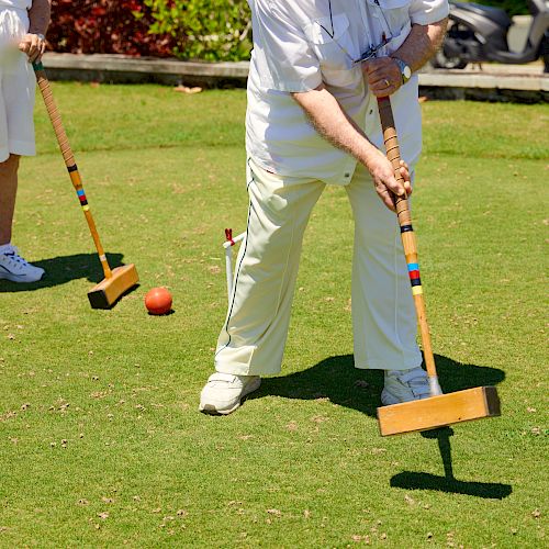 Two people are playing croquet on a grassy field. One player is about to hit the black ball with a mallet, while the other stands nearby.