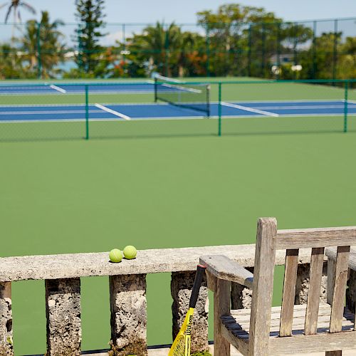 A tennis court with a few tennis balls and a racket leaning against a wooden bench, surrounded by trees and a partly cloudy sky in the background.