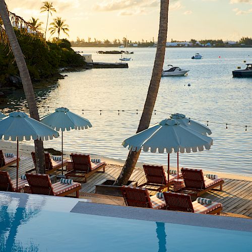 The image depicts a serene waterfront scene with lounge chairs, umbrellas, and an infinity pool overlooking the water with boats and distant greenery.